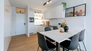Eine moderne Küche mit weißen Schränken, ein Essbereich mit einem weißen Tisch und schwarzen Stühlen sowie eine kleine Pflanze an der Wand.