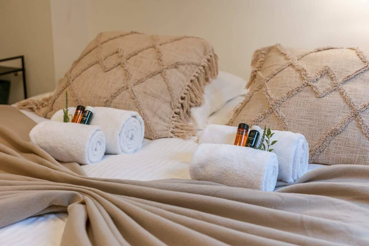 Een opgemaakt bed met opgerolde witte handdoeken, twee beige kussens en enkele toiletartikelen.