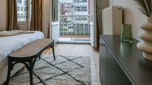 Ein Schlafzimmer mit einem Bett, einer Bank, einem Heizkörper, einem Fenster mit Vorhängen, Blick auf Gebäude und einer Vase auf einer Kommode.