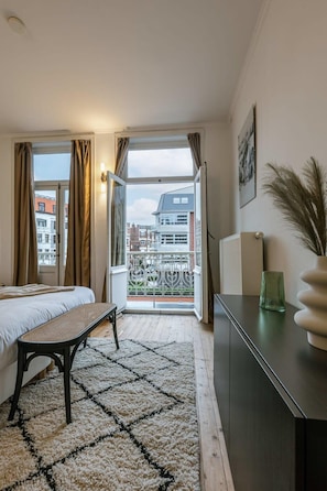 Un dormitorio con una cama, un banco, un radiador, una ventana con cortinas, vistas a edificios y un florero sobre un tocador.