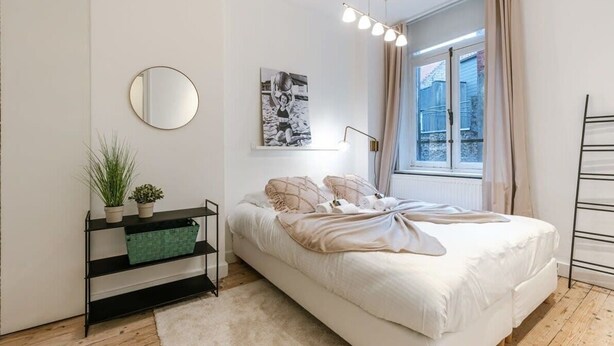 Ein Schlafzimmer mit einem Bett, einem Nachttisch mit Lampe, einem Fenster mit Vorhängen, einem Wandspiegel und einem Bild.