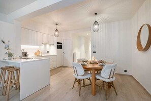 Een moderne keuken met witte kasten, een centraal eiland, houten krukken, hanglampen en een eettafel met stoelen.