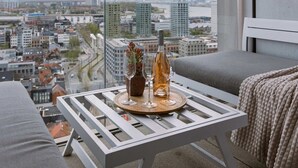 Ein Balkon mit Blick auf die Stadt, ein weißer Tisch mit einer Flasche und Gläsern sowie ein graues Sofa.