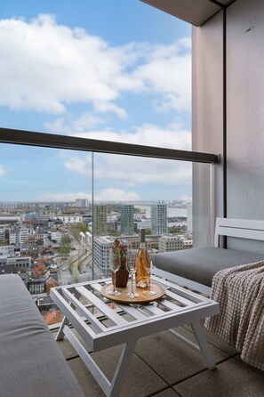Ein Balkon mit Blick auf die Stadt, ein weißer Tisch mit einer Flasche und Gläsern sowie ein graues Sofa.