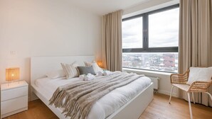 Ein Schlafzimmer mit einem großen Bett, einem Nachttisch, einem Sessel und einem Fenster mit Blick auf die Stadt.