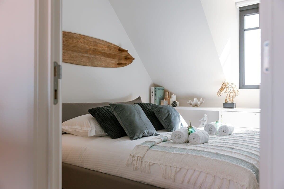 Un dormitorio con cama, almohadas, una colcha, un cuadro de madera en la pared, una ventana y una mesita de noche con libros y objetos decorativos.