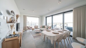 A modern living room with a dining area, large windows, and a view of the city. - Beachside apartment w/ pool & gym access (Oostende)
