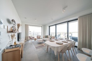 A modern living room with a dining area, large windows, and a view of the city. - Beachside apartment w/ pool & gym access (Oostende)