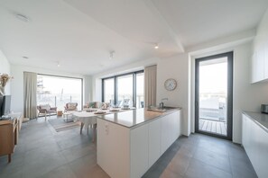 A modern kitchen with a central island, large windows, and a dining area with chairs. - Beachside apartment w/ pool & gym access (Oostende)