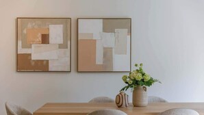 Un comedor con una mesa de madera, cuatro sillas, dos cuadros abstractos y un jarrón con flores.