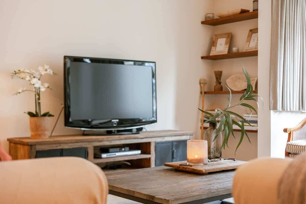 A living room with a television on a wooden stand, a small table with a candle, and shelves with decorative items.