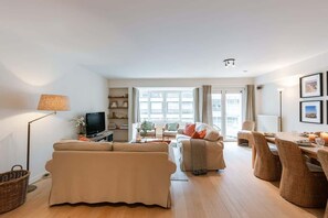 A modern living room with a sofa, a TV, a dining table with chairs, and a large window. - Beach-access apt w/ terrace & city skyline views (Knokke-Heist)