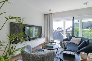 A modern living room with a flat-screen TV, a sofa, a coffee table, and a balcony with a view of buildings. - Bright 2BR apt near coastal tram w/ terrace (Knokke-Heist)
