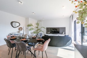 A modern living room with a dining area, a large flat-screen TV, and a large window. - Bright 2BR apt near coastal tram w/ terrace (Knokke-Heist)