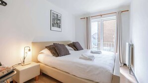 Un dormitorio con cama, mesita de noche, lámpara y ventana con cortinas.