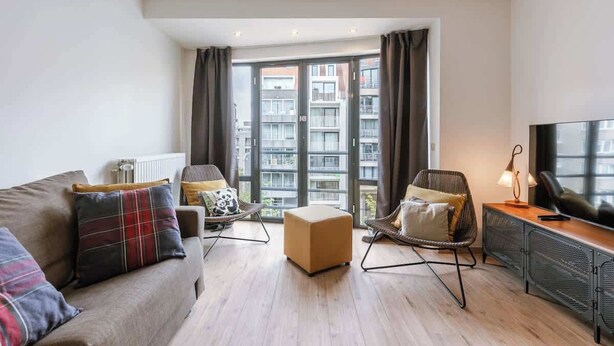 Una sala de estar moderna con sofá, reposapiés, sillas, televisor y un ventanal con cortinas.