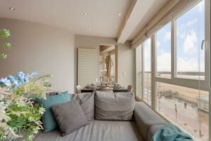 A modern living room with a sofa, a dining area, and large windows offering a view of the outside. - Seafront apt w/ open sea views, steps to beach (Knokke-Heist)