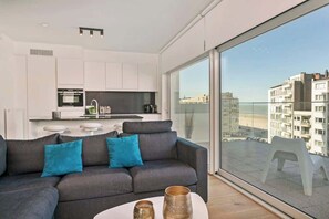 Ein modernes Wohnzimmer mit grauem Sofa, einem Couchtisch mit dekorativen Vasen, einer Küchenzeile mit weißen Schränken und einem großen Fenster mit Blick auf Gebäude.