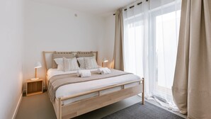 Ein modernes Schlafzimmer mit einem hölzernen Bett, Nachttischlampen, einem großen Fenster mit transparenten Vorhängen und Blick ins Freie.