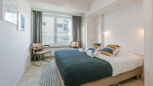 A hotel room with a bed, a chair, a window, and a view of buildings outside. - Casiola waterfront apt - garage & sea views (Oostende)