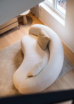 Un canapé moderne aux lignes courbes, agrémenté d’un coussin texturé et d’un coussin assorti, est placé sur un tapis de couleur claire dans une pièce dotée d’un plancher en bois et d’une fenêtre.