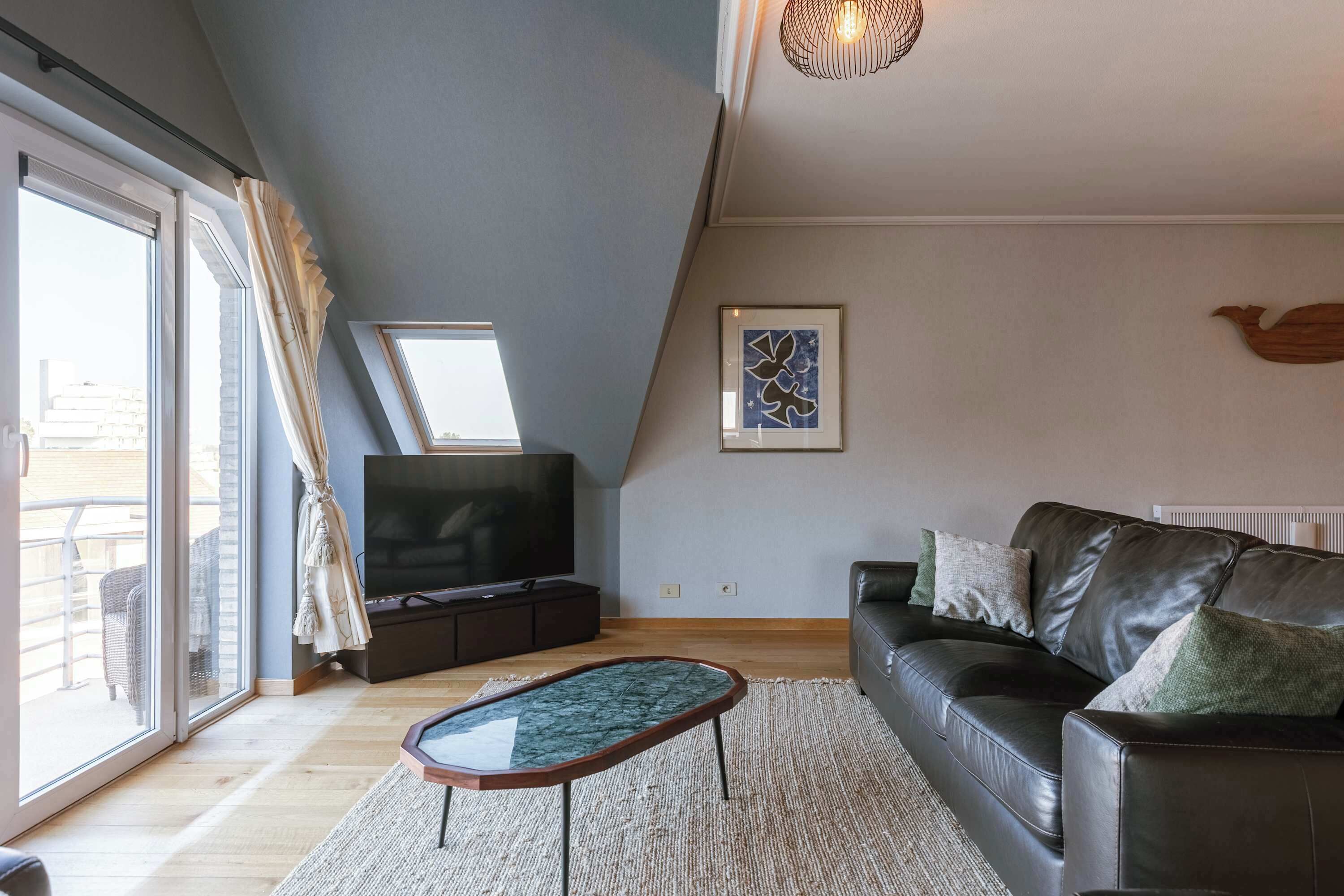 Una sala de estar moderna con una mesa de centro de vidrio, un sofá de cuero, un televisor de pantalla plana y un ventanal con cortinas.