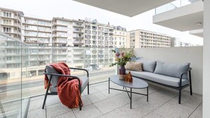 A balcony with a sofa, chair, and table, overlooking a cityscape with multi-story buildings. - Charming 2BR apartment w/ kitchen & workspace (Oostende)