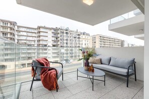Ein Balkon mit einer Couch, einem Sessel und einem Tisch, mit Blick auf eine Stadtlandschaft mit mehrstöckigen Gebäuden.