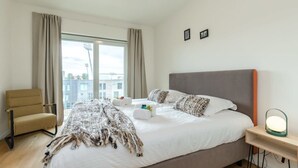 A hotel room with a large bed, a chair, a bedside table with a lamp, and a view of buildings through the window. - Charming 2BR apartment w/ kitchen & workspace (Oostende)