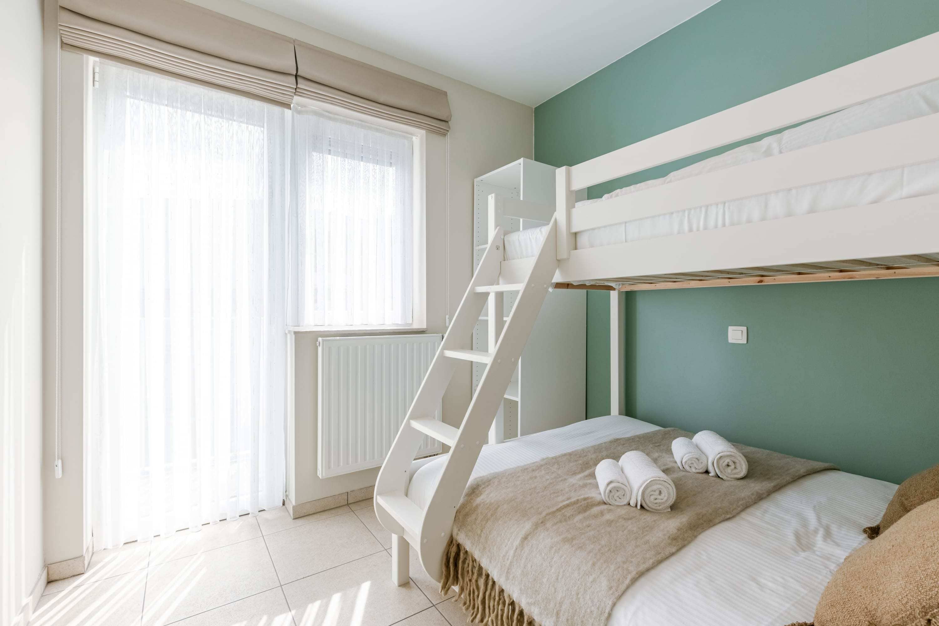 Ein Zimmer mit Etagenbetten, einer Klapptreppe, einem Fenster mit Vorhängen und einem Bett mit aufgerollten Handtüchern.