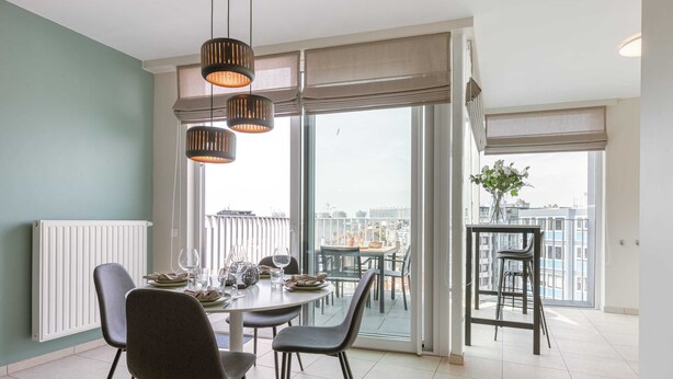 A dining area with a table set for four, a pair of chairs, and a view of the city through large windows. - Cozy 2BR apartment w/ workspace & TV (Oostende)