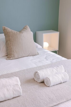 Ein ordentlich bezogenes Bett mit Kissen und aufgerollten Handtüchern, ein Nachttisch mit Lampe und eine Wand mit Lichtschalter.