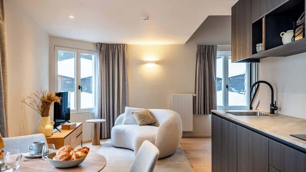 Ett modernt kök med mörka skåp, en matplats med ett bord för två personer och en mysig sittgrupp med en soffa och en fåtölj.