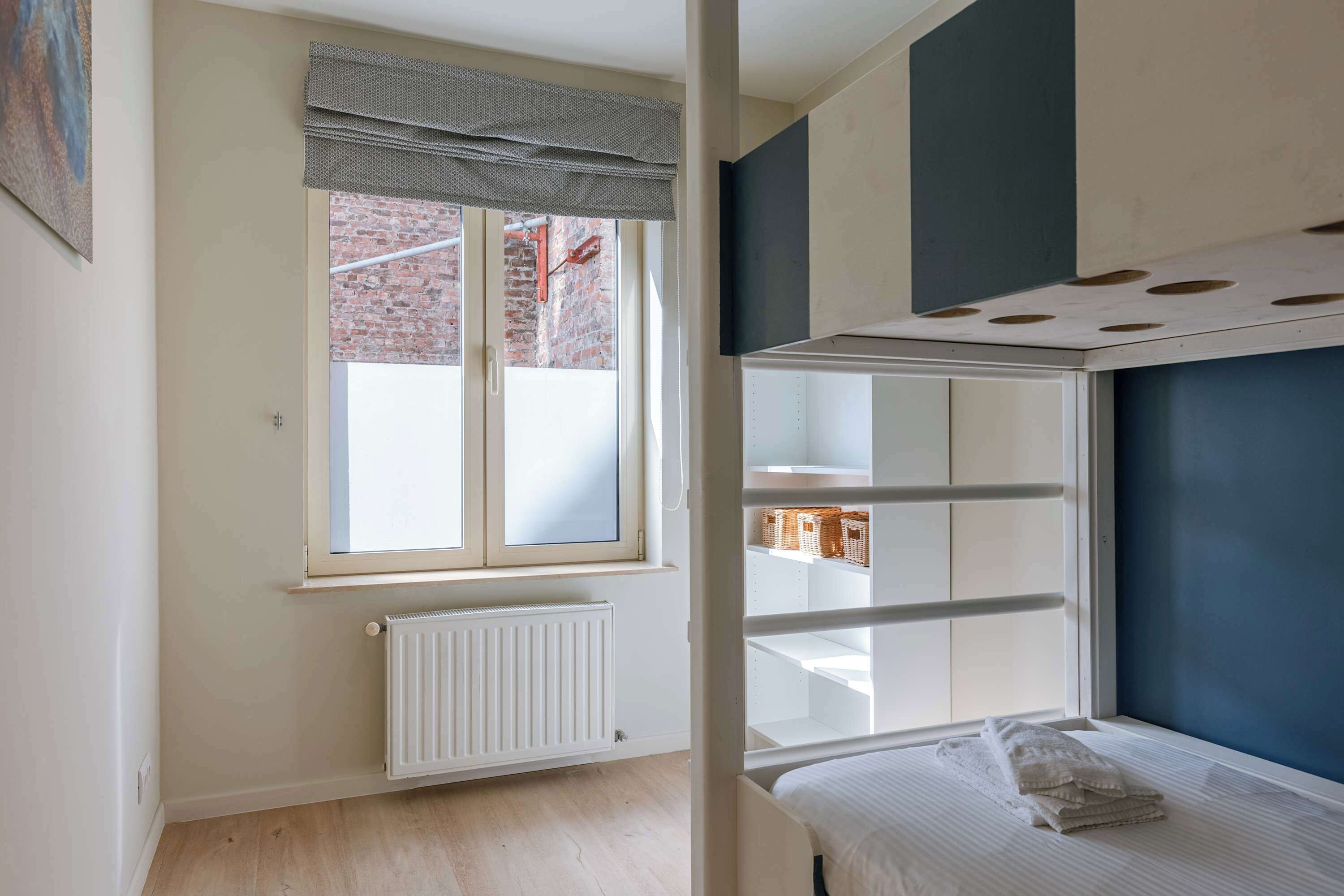 Een kamer met een stapelbed, een radiator, een raam met uitzicht op een bakstenen gebouw en een witte muur.