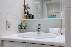 Un baño con lavamanos blanco, un espejo, una toalla y una repisa con artículos de aseo.