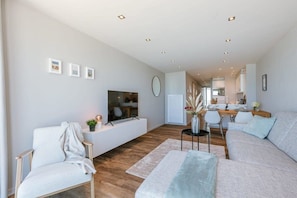 A modern living room with a sofa, a chair, a TV, and a dining area in the background.