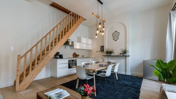 A modern kitchen with white cabinets, a wooden staircase, and a dining area with a blue rug. - Historic apartment w/ kitchen (Gent)