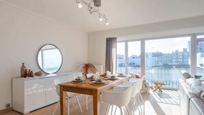 Et moderne spiseområde med et træbord dækket til fire personer, hvide stole, et hvidt skab og et stort vindue med udsigt til bygninger.