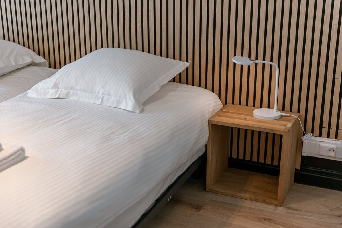 A bed with white bedding, a bedside table with a lamp, and a wooden slatted wall behind.