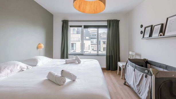 Hotelkamer met een groot bed, een ledikantje, een raam met gordijnen, een kleine tafel en een wandplank met ingelijste foto's.