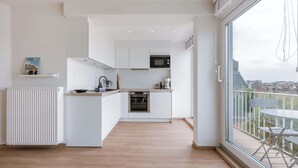 Een moderne keuken met witte kasten, een inbouwoven en een magnetron. Er is een eethoek met een tafel en stoelen bij een groot raam.