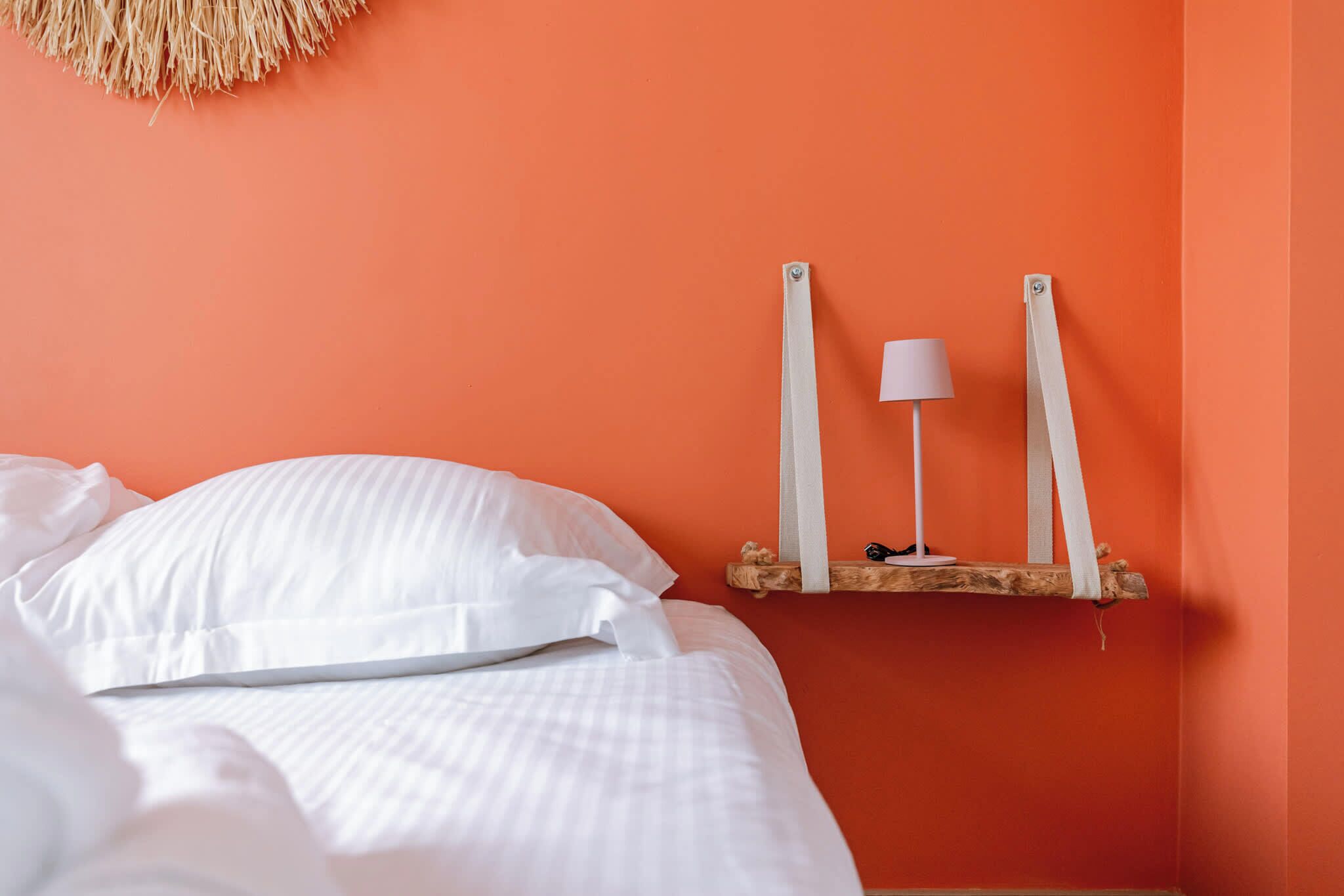 Un lit avec une literie blanche, une lampe blanche posée sur une étagère en bois et un mur orange.