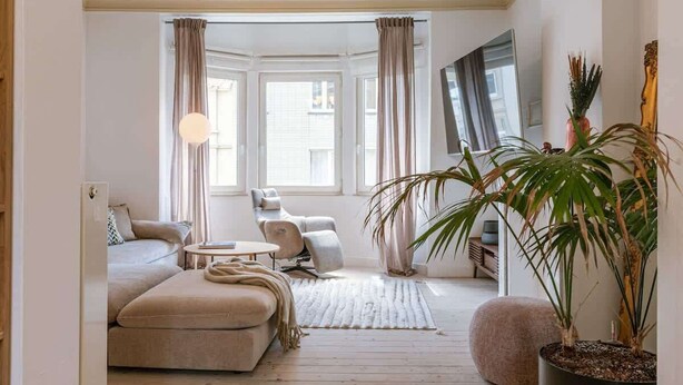 Ein Wohnzimmer mit Sofa, Sessel und Couchtisch. Ein großes Fenster mit Vorhängen, ein Blumentopf und ein Spiegel an der Wand sind ebenfalls zu sehen.