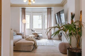Una sala de estar con sofá, sillón y mesa de centro. Hay un ventanal con cortinas, una planta en maceta y un espejo en la pared.