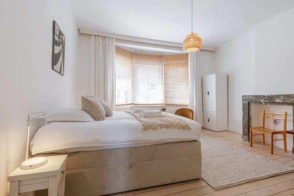 Un dormitorio moderno con una cama grande, mesita de noche, lámpara y vistas a un paisaje urbano a través de la ventana.