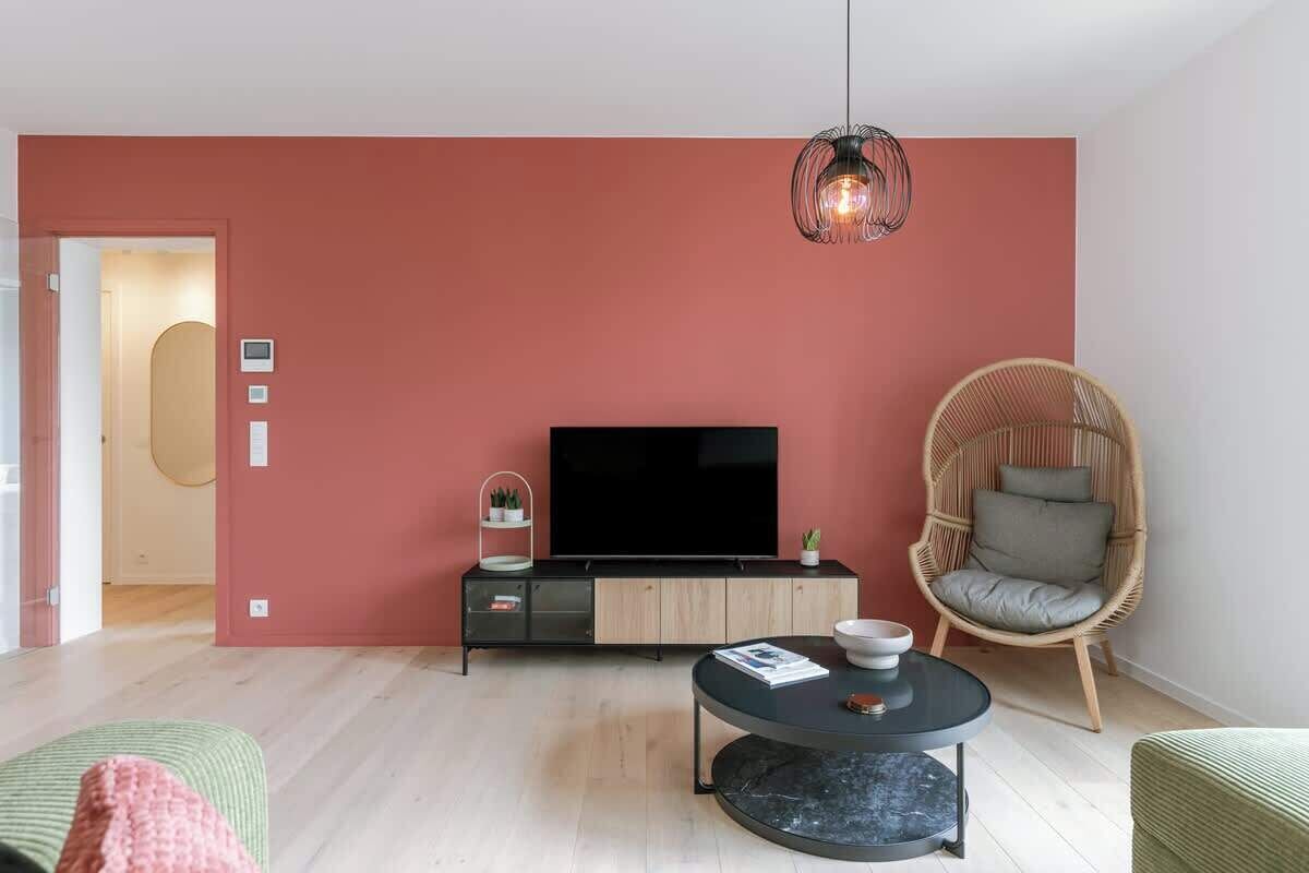 Ein modernes Wohnzimmer mit Flachbildfernseher, rundem Couchtisch, Korbstuhl und einer roten Akzentwand.
