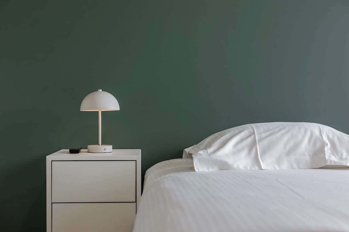 Una cama con ropa blanca, una mesita de noche blanca con una lámpara y una pared verde.