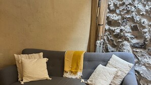 A grey sofa with white and yellow cushions, a round wooden table, and a stone wall. - Holiday Home 'Gite Les 4 Saisons' with Mountain View, Private Terrace and Wi-Fi (Val de Chaise)