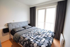 A bedroom with a bed, a nightstand, a window with curtains, and a radiator. - Waterfront promenade apt w/ beach access (Middelkerke)