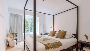 A four-poster bed with a white and beige bedspread, a bedside table with a lamp, and a view of a balcony with curtains. - Family-ready apt w/ game console - heart of Ostend (Oostende)